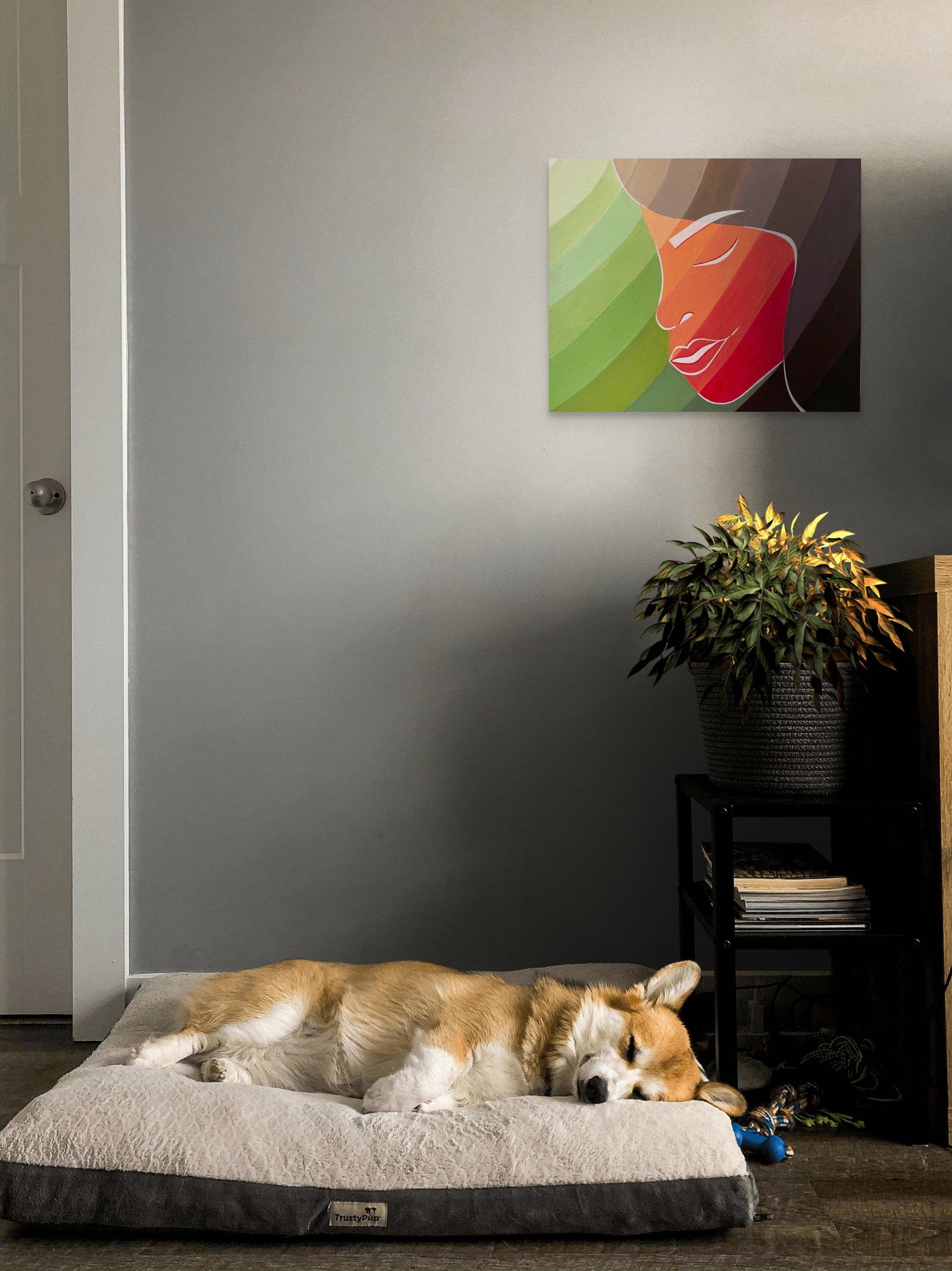 Painting of a young woman is hanging in a lounge above a dog bed with a Corgi.